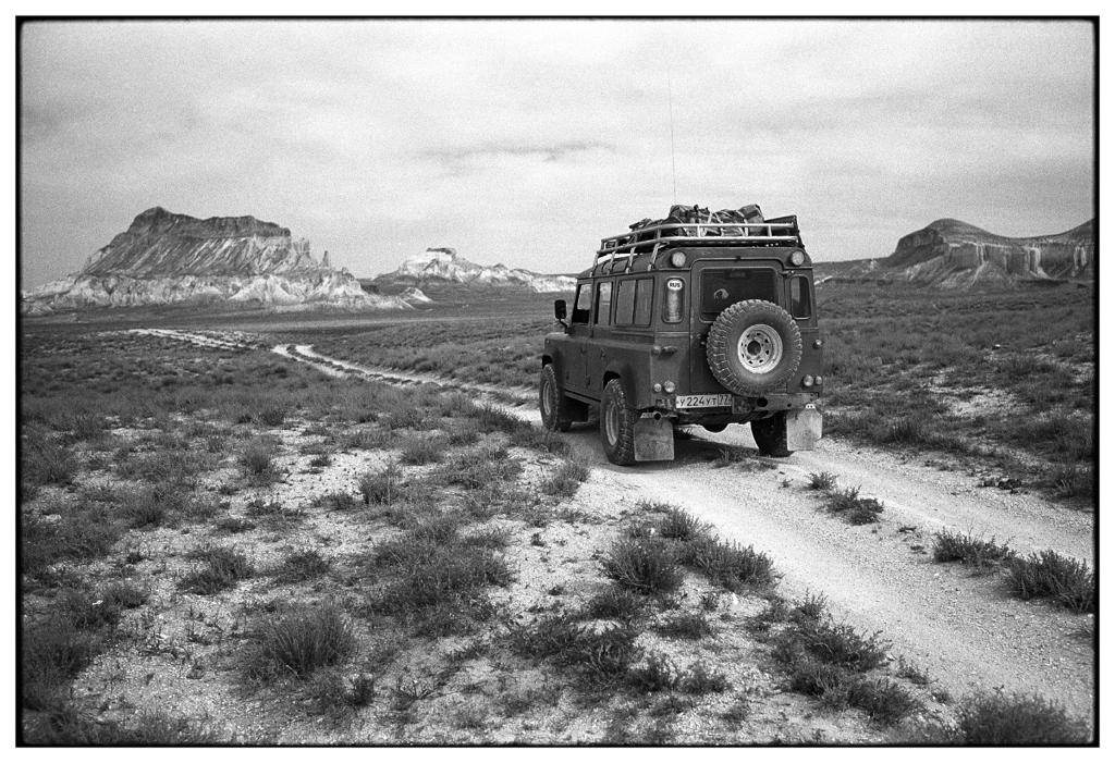 Black and white photograph of a Land Rover Defender driving on a dirt road towards dramatic desert mountains